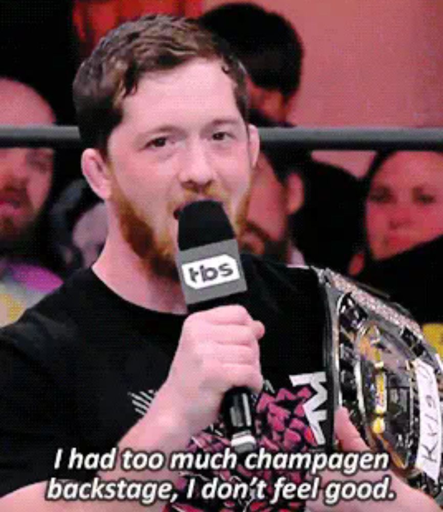 still from aew dynamite showing kyle o'reilly talking into a mic. he's saying 'I had too much champagen backstage, I dont feel good' and holding a title belt. the belt has a piece of tape on it with 'kyle' crudely written in marker. the e in kyle is backwards