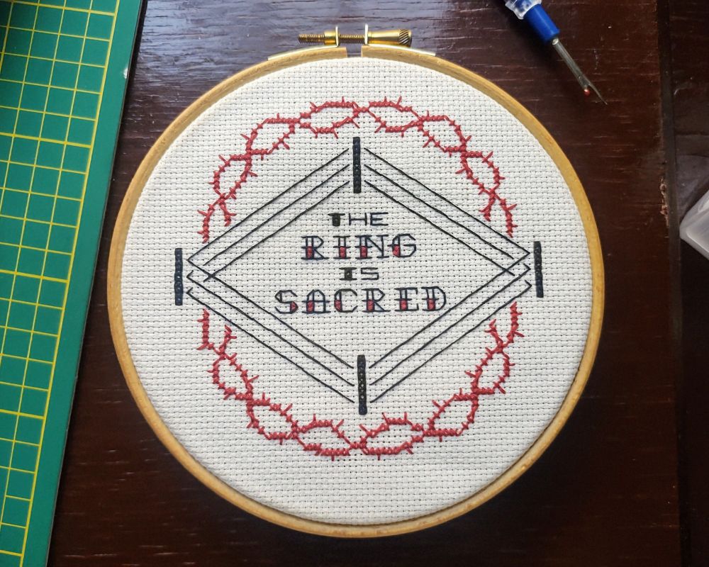 a black and red cross stitch pattern on white cloth - the words 'the ring is sacred' in the center of a wrestling ring, surrounded by a circle of red barbed thorns