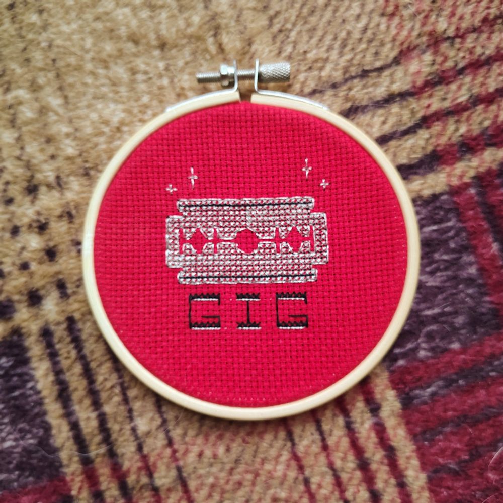 a cross stitch hoop. the cross stitch is a metallic silver razor blade on red cloth, below the word 'GIG' 