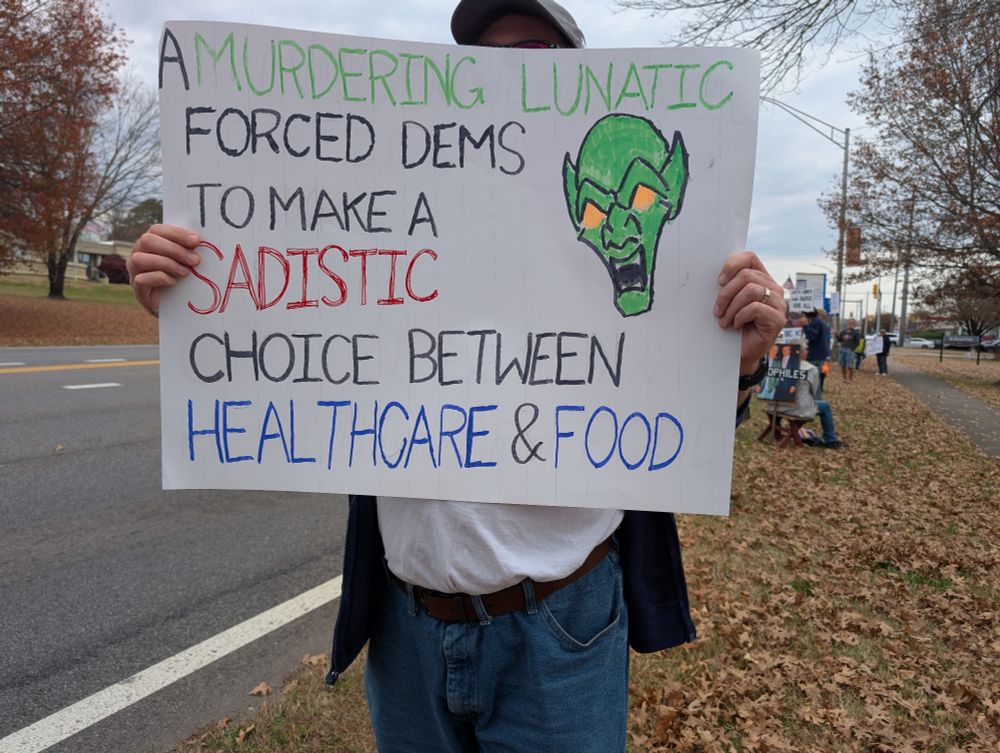 Protester holding a sign that has a hand drawn green monster face with yellow eyes, and an open mouth with pointy white teeth, on the right with the words, "a murdering lunatic forced dems to make a sadistic choice between healthcare & food" surrounding the face and taking up the rest of the area on the white poster board.