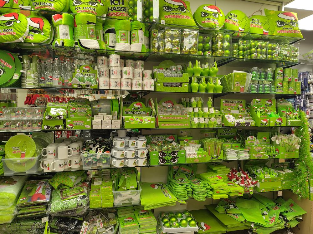 jede menge Grinch nachahmende Produkte in einem ladenregal. auf dem steht "ich hasse menschen" es gibt Pappteller, Socken, Stifte, Tassen, Handschuhe, Weihnachtsbaum kugeln und viel mehr