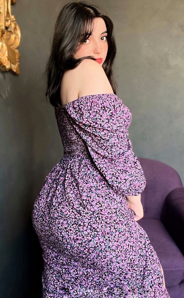 A woman with dark hair wearing an off-shoulder, fitted dress with a black and purple floral pattern is standing and posing with her body turned slightly to the side, glancing over her shoulder at the camera. Behind her is a purple armchair, and a golden decorative element is partially visible on the textured, neutral-colored wall in the background.