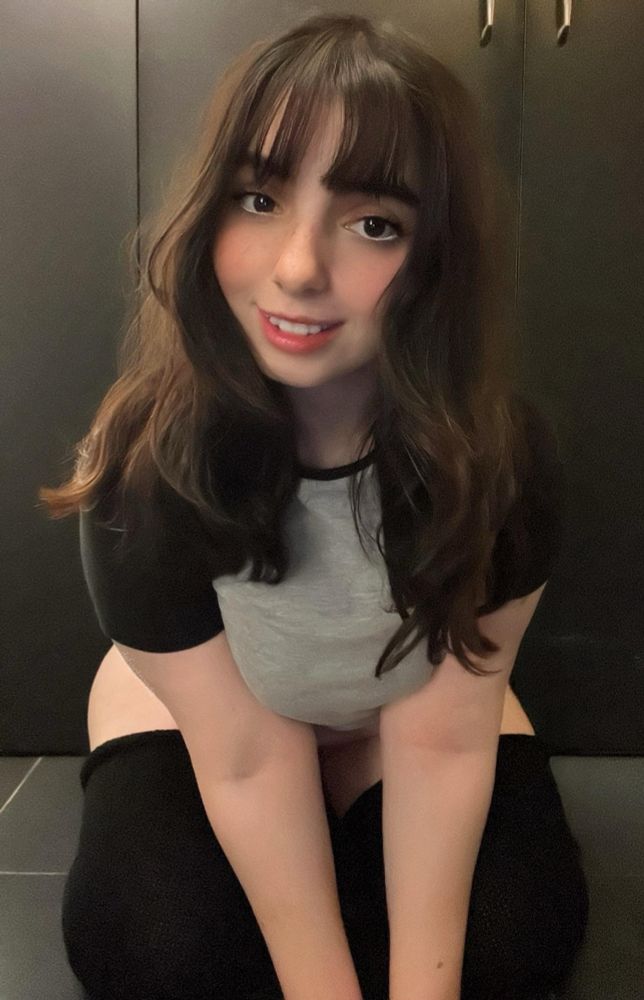 A young woman with long, wavy dark brown hair and bangs is smiling at the camera. She is wearing a short-sleeved, black and gray top and black thigh-high socks, seated on the floor and leaning slightly forward. The background features dark cabinets.