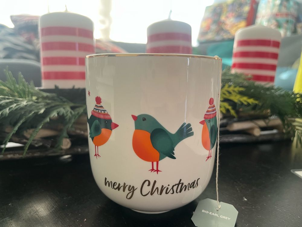 Vor einem Adventsgesteck mit Kerzen steht Eine Tasse auf der 3 weihnachtliche Vögel zu sehen sind und auf der in Gold Merry Christmas steht. Ein Teebeutel hängt heraus. Im Hintergrund sieht man Adventskalender auf einer Couch stehen.