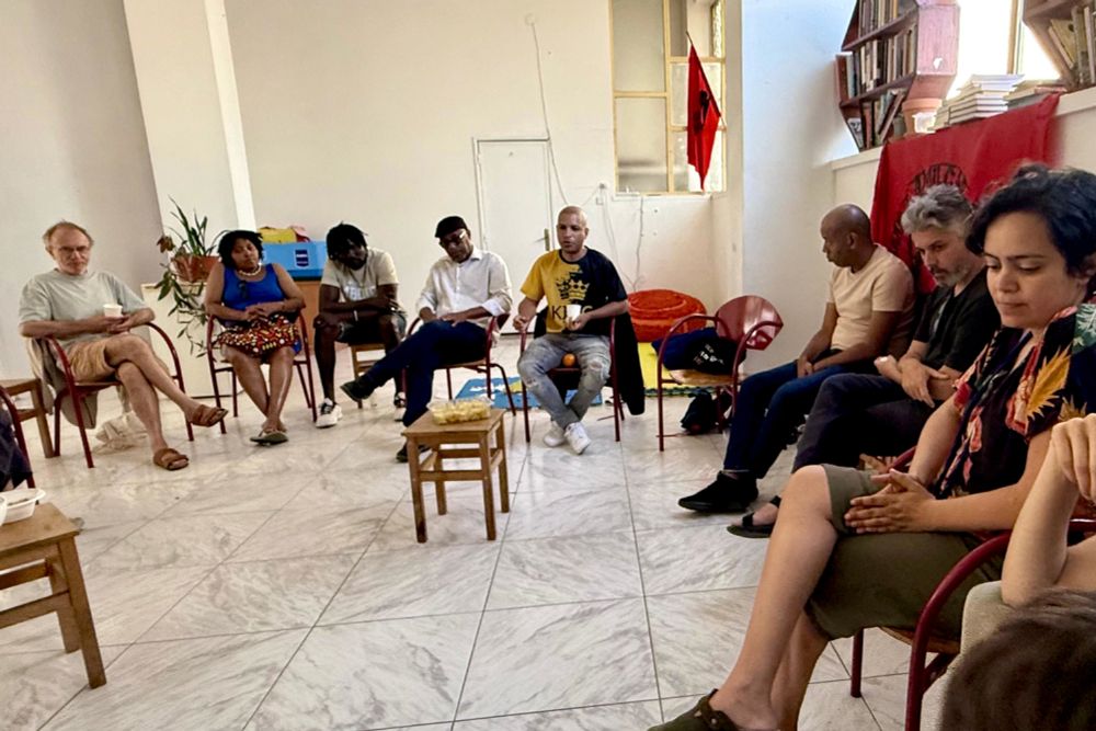 Conversation with Fernando Moreira in the the Mbongi_67 Cultural Space. People are seated in a circle. In the middle, there's a wood bench with a tray of chips (crisps)