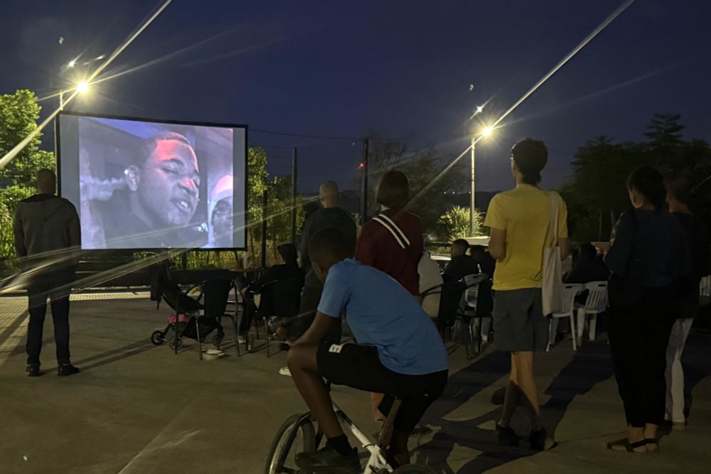 Screening in the Bairro do Zambujal football courtyard. It's night time and there are some people seating on plastic chairs, some people standing, and a boy watching from a bicycle