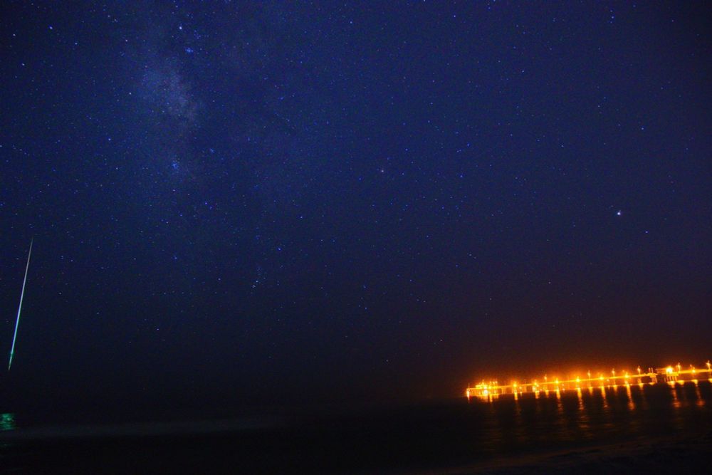 Milky Way over the ocean and pier, featuring a meteor photobomber.