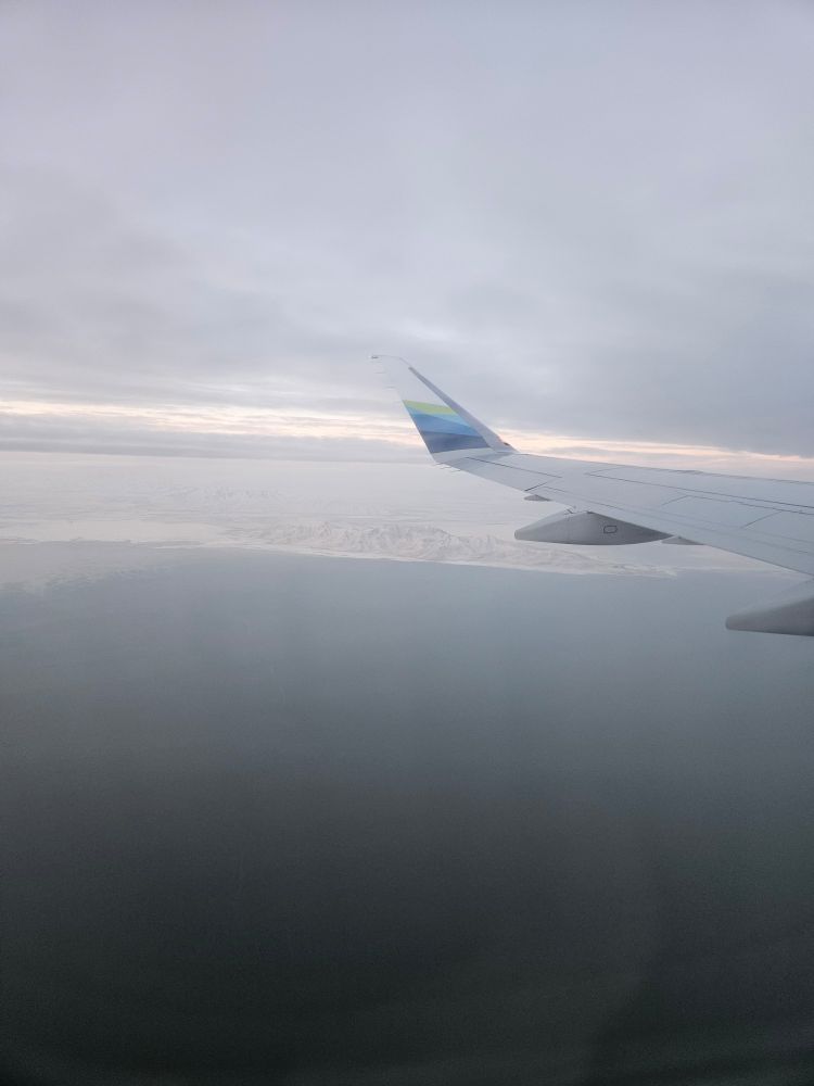 Pretty sure the mountain at the edge of the lake is Stansbury Island. This is the second largest island of the lake and has a significant land bridge connecting it to the surrounding lands. Evaporation ponds now flank its west side, which are part of the mineral extraction process.

Photo description: A view from an airplane showing the airplane wing and a cloudy morning over the Great Salt Lake. The water is blue and the land where it meets is snowy and pale. A sliver of silvery-rose sunrise color is still visible in the horizon, even in the pale morning light.