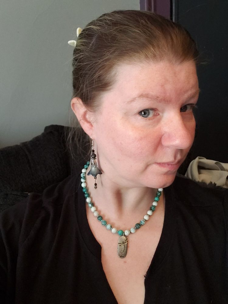 A white person wearing a black shirt, a pair of black and silver earrings in the shape of an inverted flower, and a green and white beaded necklace with a silver dragonfly pendant. 