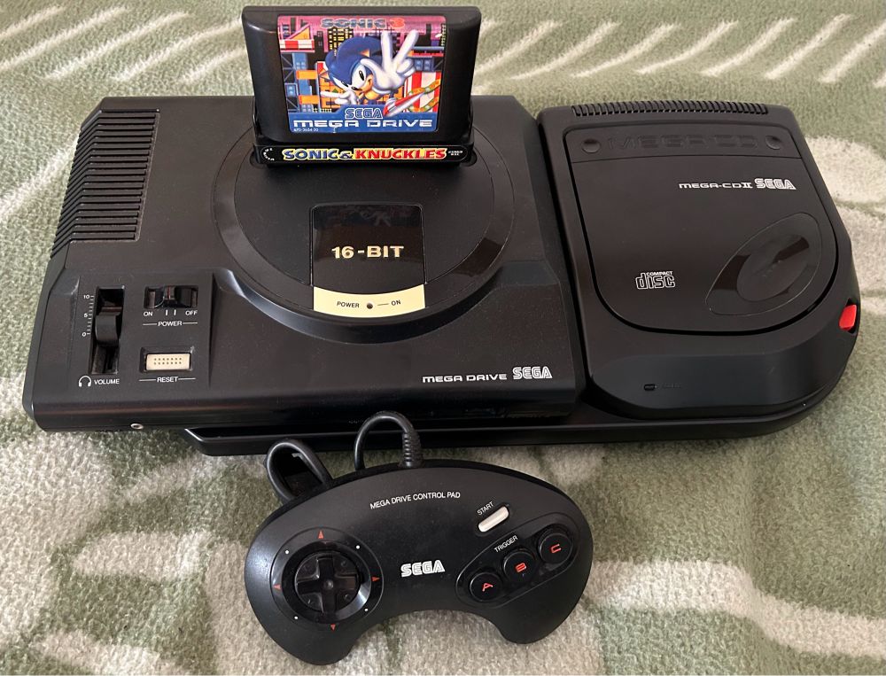 Sega MegaDrive Model 1 With Sega Mega CD 2 Add-on attached to it, as well as a MegaDrive Control Pad in front of it and a copy of Sonic 3&K plugged into the cartridge slot.