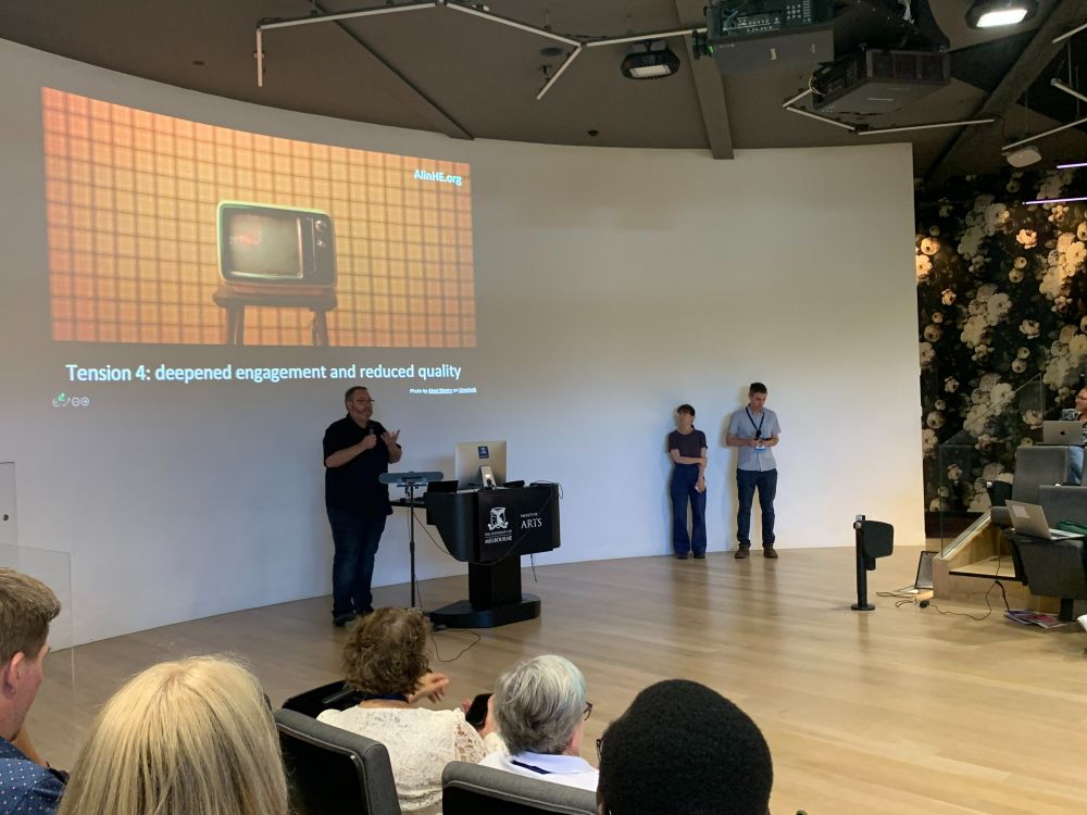 Michael Henderson presenting below a photo of an old-style TV with the text "Tension 4: deepened engagement and reduced quality". Margaret Bearman and Tim Fawns are standing to the right. Some members of the crowd are visible. 