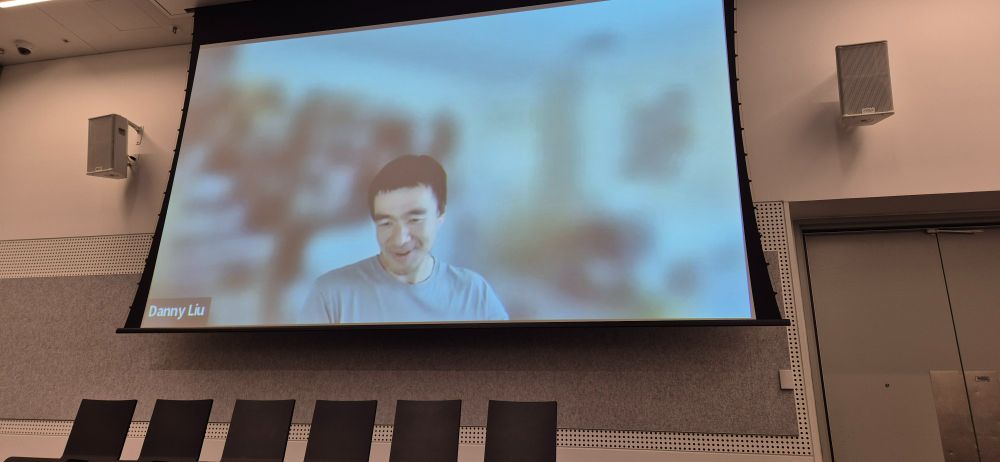 Danny Liu against blurred Zoom background, projected on a conference wall.