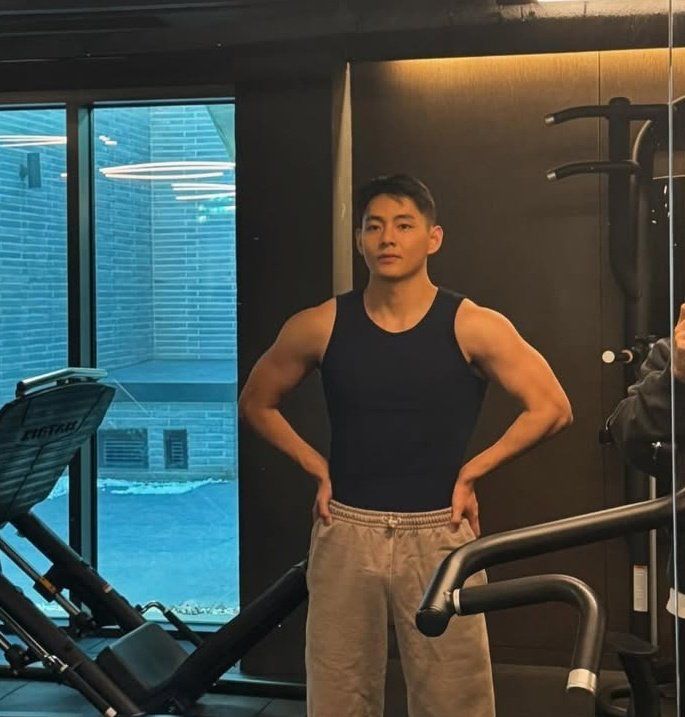 V of BTS in the gym wearing black tank top and gray sweatpants looking buff. 