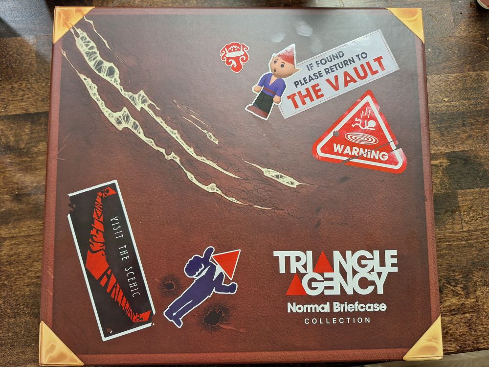 A box that says Triangle Agency Normal Briefcase Collection. The cover is made to look like a distressed briefcase with stickers of a gnome and a few unseemly holes on it.
