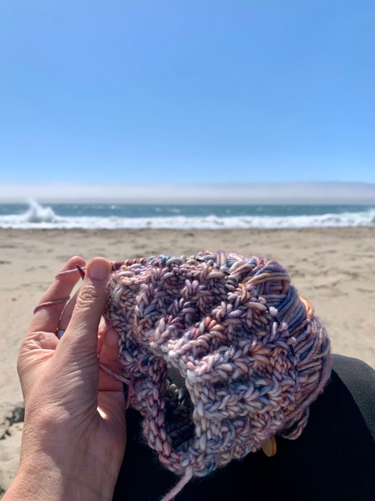Pacific Ocean, fog bank, and sky in the background. Multi color (blues, pinks, neutrals) piece of knitting and yarn with a hand and knitting needles. 
GINEVRA pattern by Wanded
Malabrigo Mecha in Hannah 
