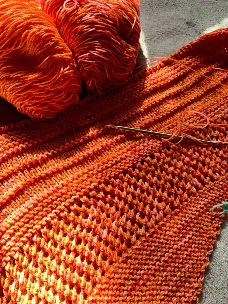 Hand knit shawl in progress with partial lace pattern; made with two different balls of yarn. One ball is orange, and the other variegated colors such as pink, red, orange and mustard, both shown in the picture. One strand from each ball of yarn used, held together, to make the shawl. One knitting needle can also be seen. 
