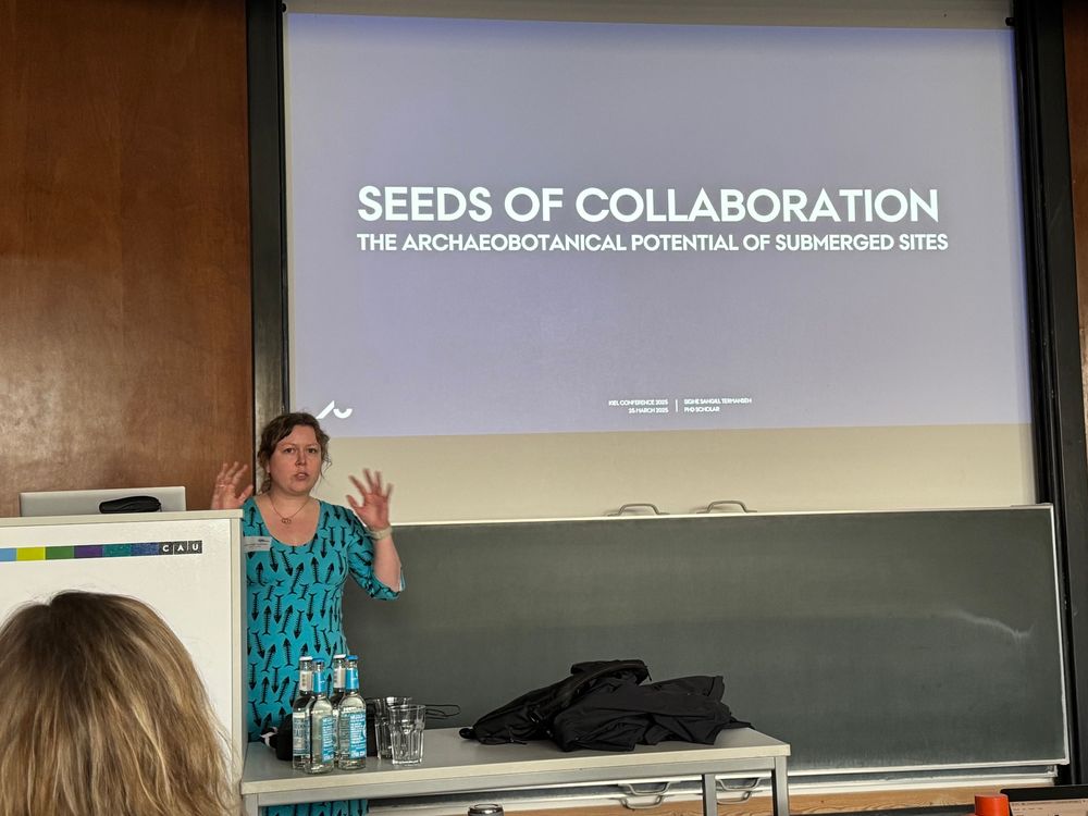 Archaeobotanist in blue dress with fish skeletons gesticulates wildly while trying to convince other people of the value of studying plants in a conference room. The slide in the background says “Seeds of collaboration. The archaeobotanical potential of submerged sites”. 