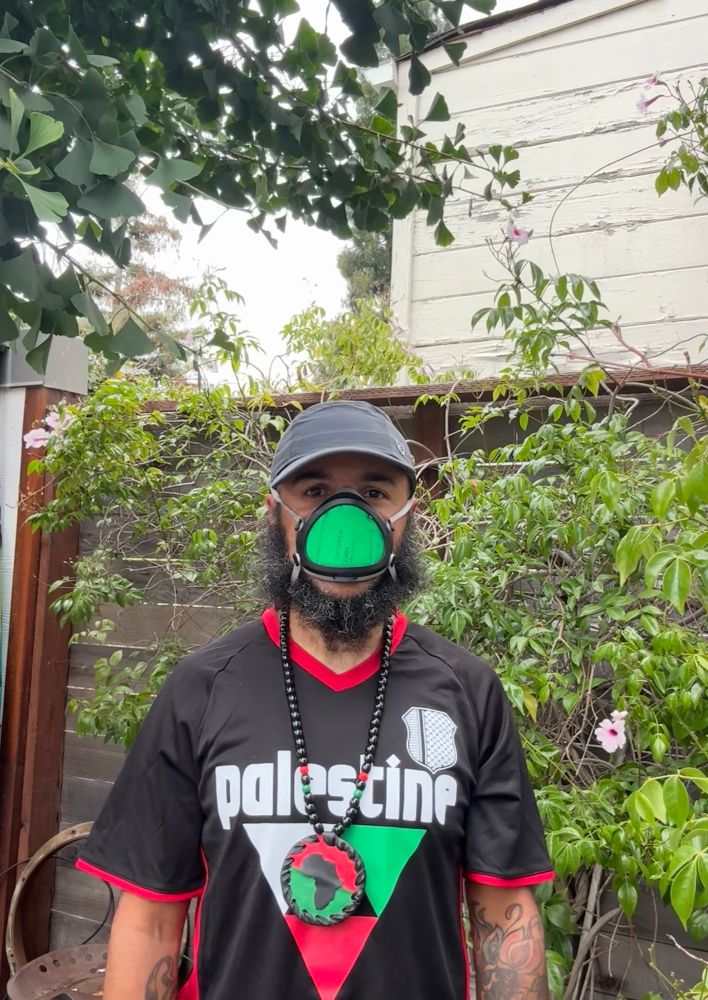 background is a fence and plant life, trees and such.  standing in the center is a light complected black man wearing a black hat, black/green mask, a black jersey that says “palestine” with the same color combo red/black/green african medallion necklace