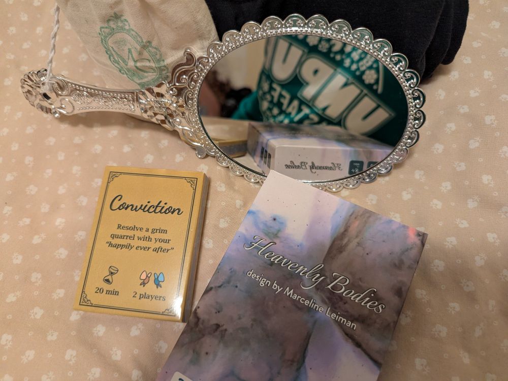 "Conviction" and "Heavenly Bodies" on a fabric spread, with a hand mirror from "Gorgon's Garden" reflecting them for the camera