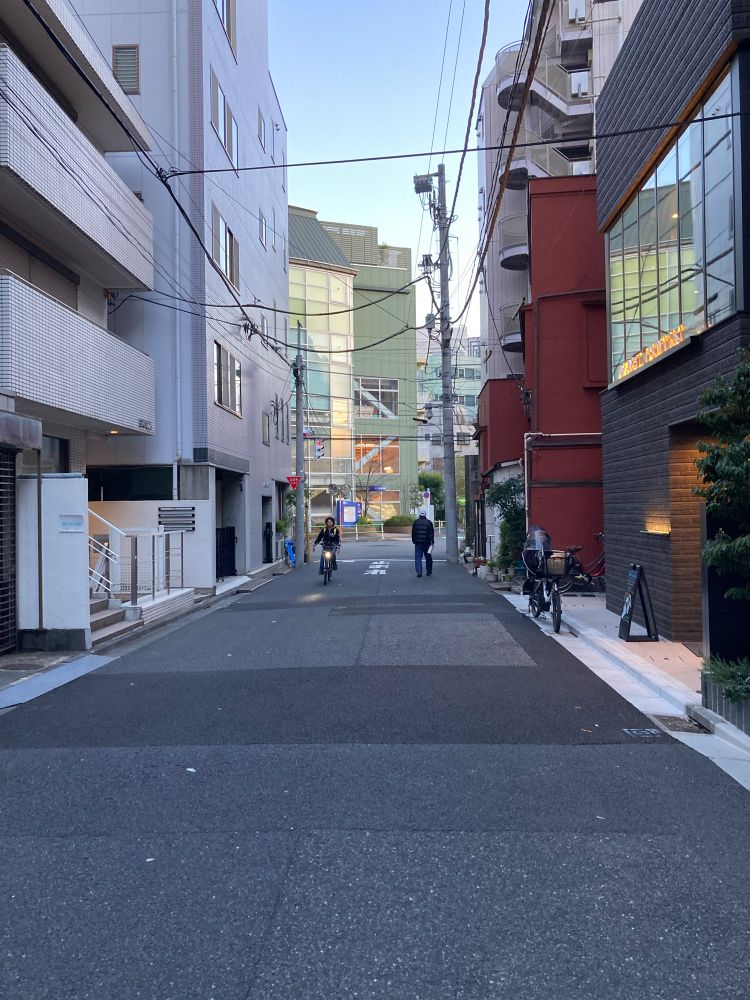 Eine Nebenstrasse mitten in Herzen von Tokyo, im Winter. Es ist kein Auto zu sehen, weder parkend noch fahrend. Aber ein Fussgänger sowie ein Radfahrender. Am Strassenrand ist ein weiteres Fahrrad geparkt.