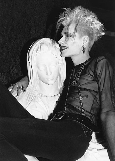 John Koviak looking like a goth lesbian (spiky hair and sideshave, black lipstick and eyeliner, cross earrings and necklace, sheer black shirt, black jeans) sitting in the lap of a statue of the Pietà