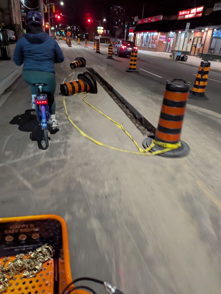 A roadway with traffic cones and caution tape; to the right of the left-most land, deep trenches have been cut to allow concrete barriers to be installed, permanently and physically separating the bike lane from traffic.