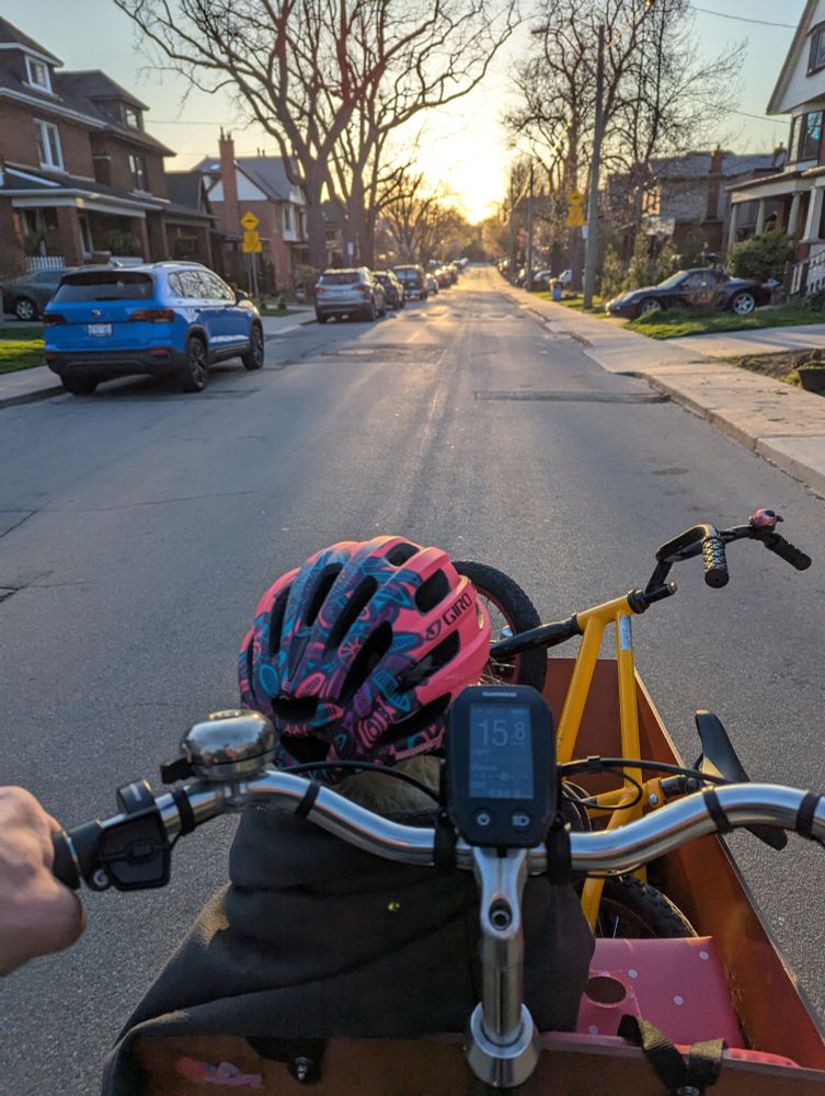 A young child sits in the front of a Dutch style cargo bike, snuggling in a blanket, with their small child's bike beside them. in the background, the sun sets behind trees at the end of a quiet residential street. Taken from the perspective of the cargo bike pilot.