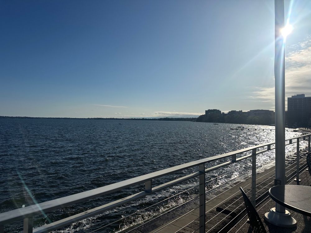 A picture of a lake. The sun is shining. There is a railing in the foreground of the photo. 