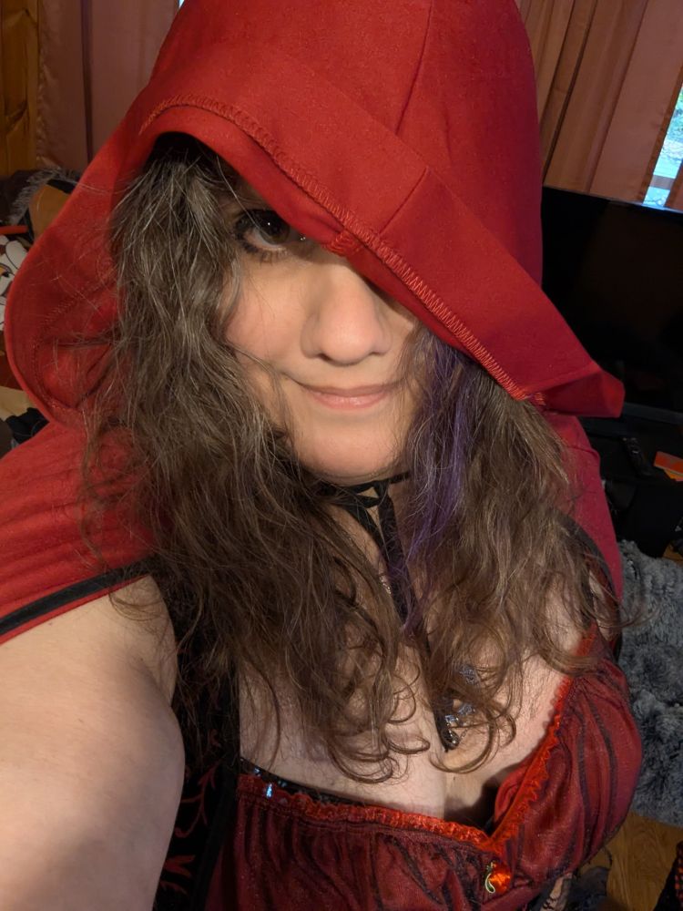 A lady in a red, hooded cloak and low-cut dress as her Halloween costume, one eye covered by the hood.