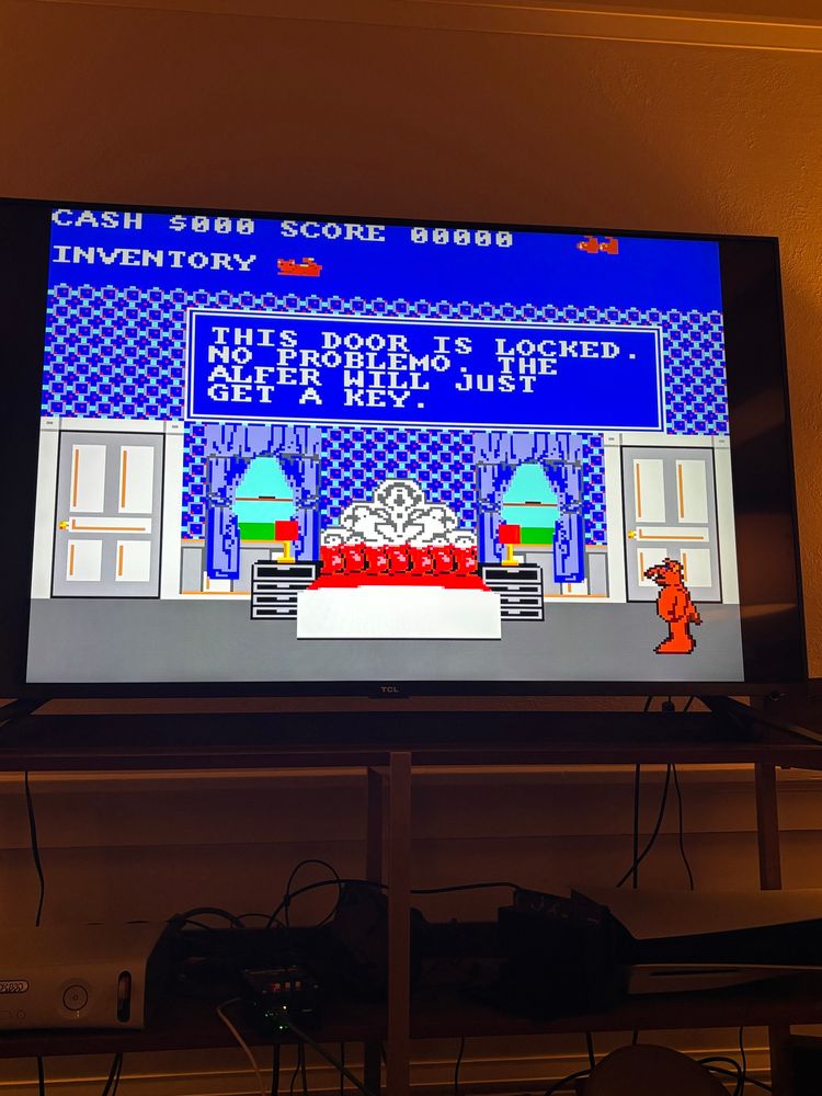 A photo of a tv with the sega master system game “alf”. Alf stands in a blue bedroom with a red bed between two blue windows and two white doors. A dialogue box reads  “THIS DOOR IS LOCKED. NO PROBLEMO. THE ALFER WILL JUST GET A KEY”