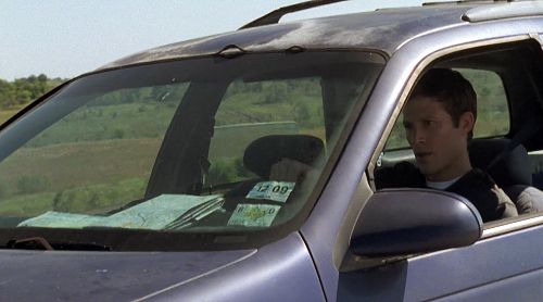 Zach Gilford as Matt Saracen on Friday Night Lights in a grey car driving on a sunny day, a map on his dashboard