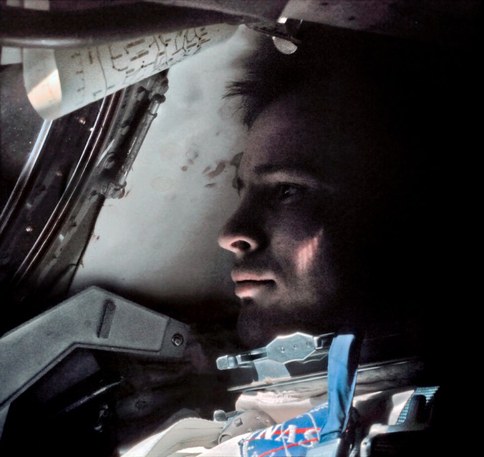 A photo of the side profile of an astronaut inside his capsule. He's looking to the left. He's wearing a space suit (without the helmet) and the 1960s version of the NASA logo is visible on a patch on the suit. He's next to the wall of the capsule and you can see a little bit of the window he's looking out of. Above his head is a rolled up paper that's a star chart of some sort. 