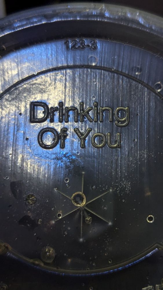 The image shows the textured surface of a black drink lid. In the center of the lid, the phrase "Drinking Of You" is embossed in bold, uppercase letters. Surrounding the text, faint circular grooves and small droplets of liquid are visible, reflecting light. Above the phrase, the number "123-3" is also embossed. The overall lighting casts a bluish tint on the lid, creating a slightly reflective effect.