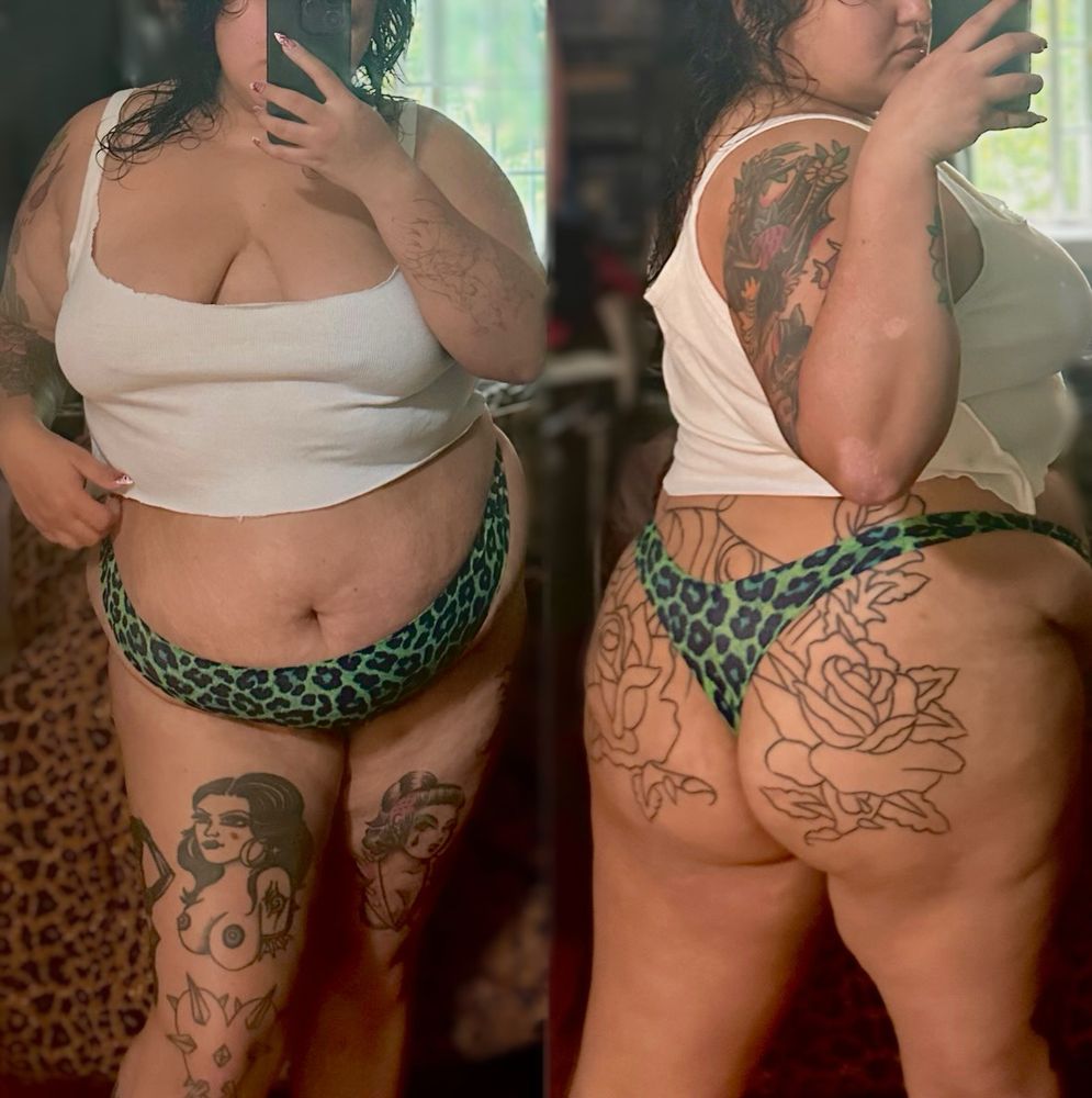 Two mirror selfies of Mistress Dahlia combined into one sexy photo. Both sides of the image showing Mistress Dahlia with wet hair holding her phone near her face to take the selfie, her hair wet and dripping just like her pussy. She’s in the tiniest cut up white tank top, a green leopard print thong that hugs her tummy and juicy tattooed ass perfectly. You see her beautiful brown skin glowing, covered in American traditional color tattoos of pinups, roses, and a fruit bat all over her body. She is the whole package of curvy Latina excellence. You’ll want to book her now to experience it yourself. 