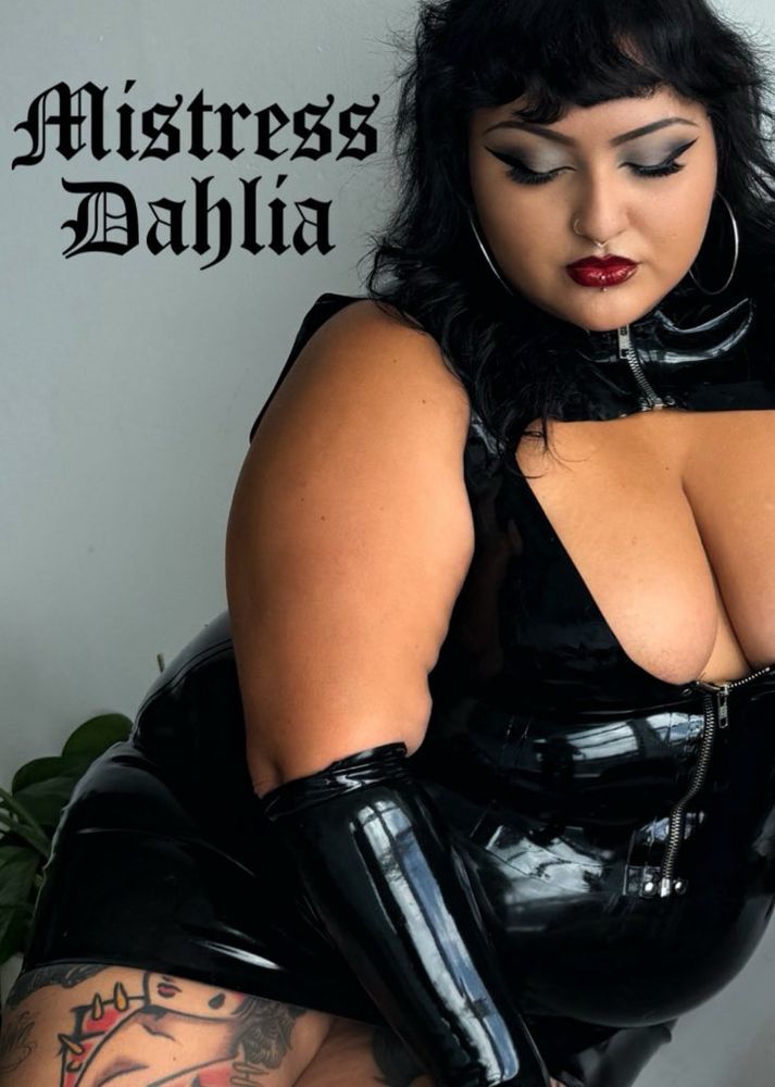 An image of Mistress Dahlia in all black latex in front of a grey background with the text “Mistress Dahlia” in old English in the back. She is looking down with her long lashes and smokey grey eyeshadowed eyes, a red lip, and a low plunge latex top that shows off her bust. Latex gloves come up to her elbow, her latex skirt short as always and revealing her thigh tattoos. 