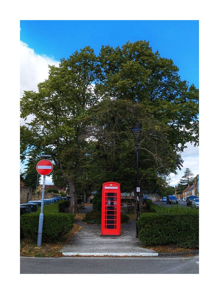 A colour photo of a bright red phone box at one end of a small urban green square, standing between a red "no entry" street sign and a black street lamp, against a backdrop of trees and blue sky.