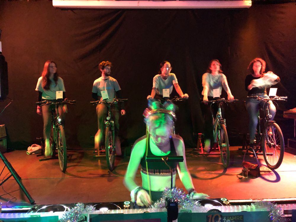 Fünf Fahrräder stehen auf der Bühne und erzeugen Strom über einen Generator am Hinterrad. Der Strom wird genutzt um die Sound Anlage und das Display, den DJ Pult und etwas Licht zu betreiben. Das System ist autark und kann ganz ohne Stromanschluss vom öffentlichen Netz auskommen. Im Vordergrund steht eine DJane an ihrem Pult. Dahinter auf der Bühne sind fünf Menschen auf dem Fahrrad am  radeln, um die Energie für die Disco zu erzeugen.