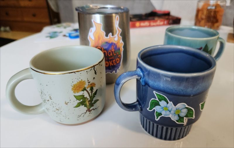 A photo of some mugs that also have botanical imagery - a dandelion, a dogwood flower. In the background is a tumblr with a flaming planet and "Chat, are we Cooked?" on it
