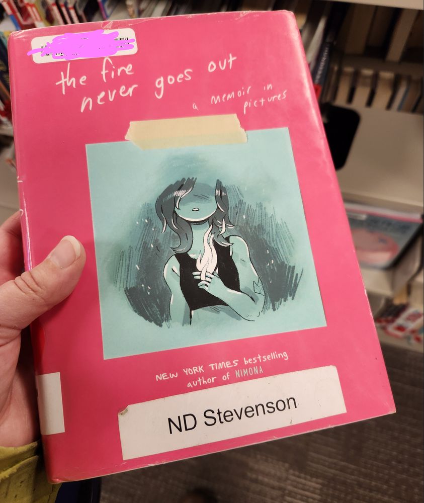 A photo of jam's hand holding a copy of 'the fire never goes out - a memoir in pictures'. A handmade label has updated the author's name to 'ND Stevenson' :)