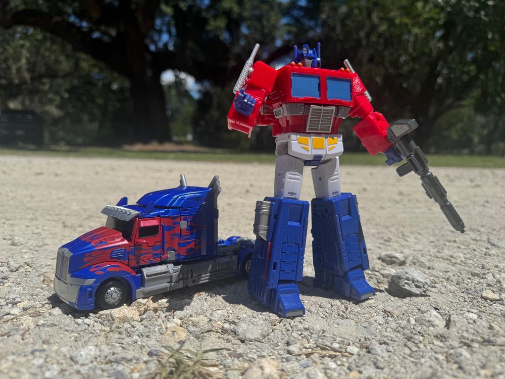 AoE Optimus in truck mode (left) and 86 Optimus (right) standing on a rocky dirt road.