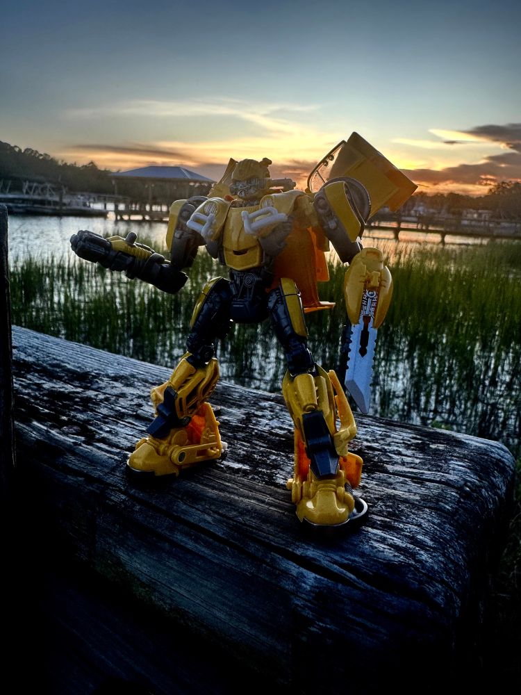 Bumblebee in robot mode in front of a sunset with weapons in hand.