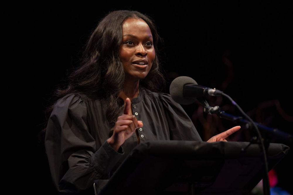 Yetide Badaki stands on the stage in front of a microphone reading a story.