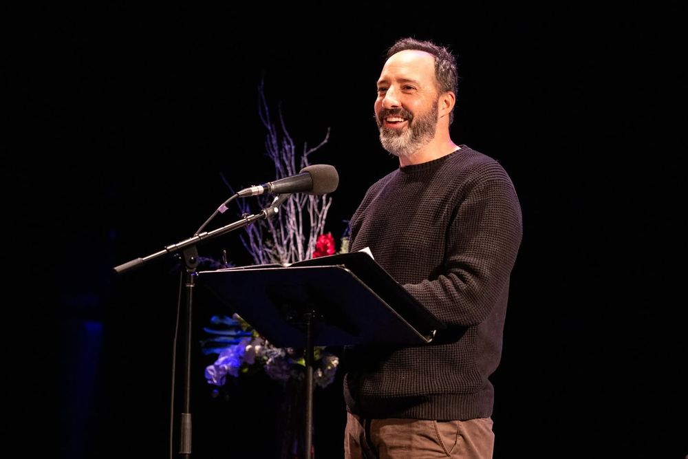 Tony Hale stands on the stage in front of a microphone reading a story.