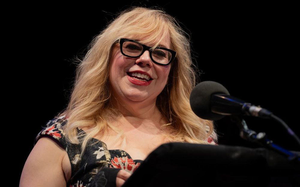 Kirsten Vangsness stands on the stage in front of a microphone reading a story.