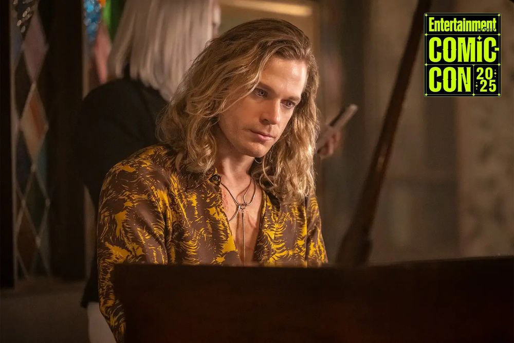 Lestat from a comic con sneak peek of The Vampire Lestat. He’s sitting at a table in an open yellow and brown shirt. 