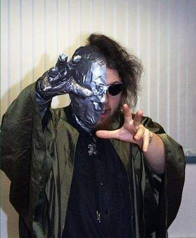 Me, age 22ish.  The entire right side of my head is covered in duct tape.  I am wearing a pair of sunglasses missing the right lens.  The frames blend in with the tape.  My right hand is also wrapped thusly.  Also a green choir gown is worn.  I am gesturing toward the camera, almost menacingly.