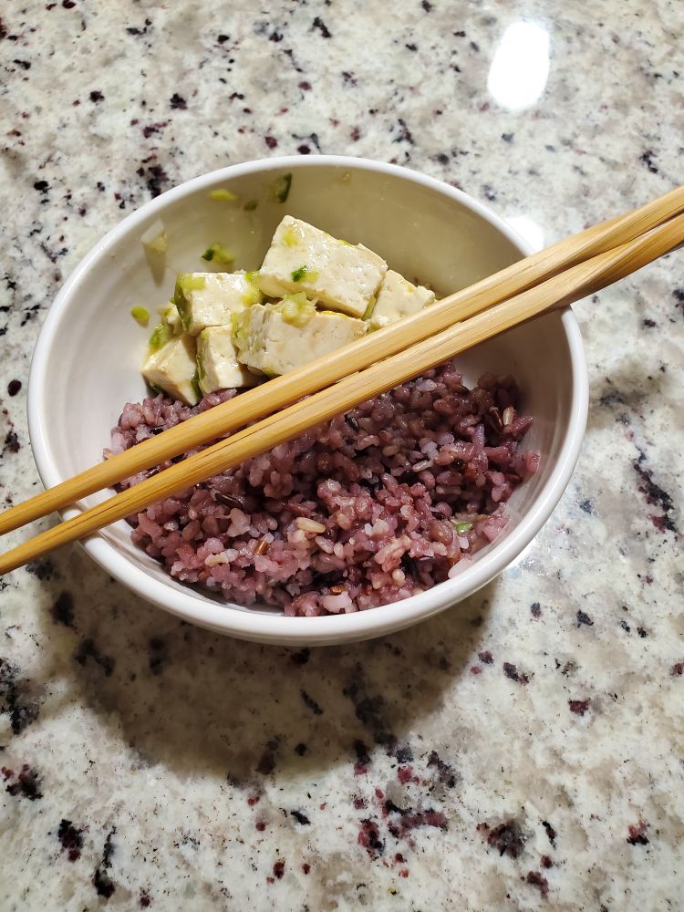 Close-up photo of a bowl of dressed tofu with mixed grain rice and a pair of wooden chopsticks laid across the top