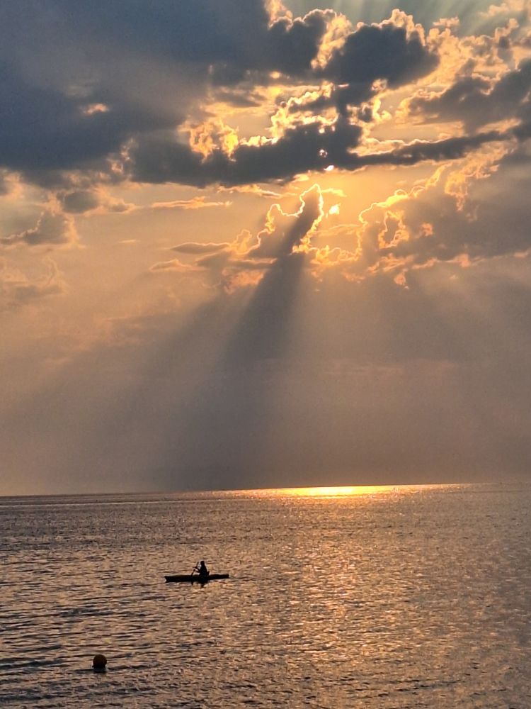 A pretty amazing sunset behind clouds, with rays casting all about the water below, with a kayaker in the foreground 