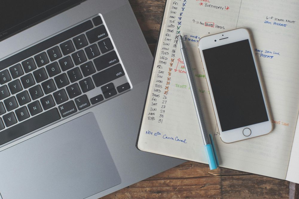A laptop and a smartphone sitting next to each other on a table, with the latter lying on top of a day planner full of handwritten notes.
