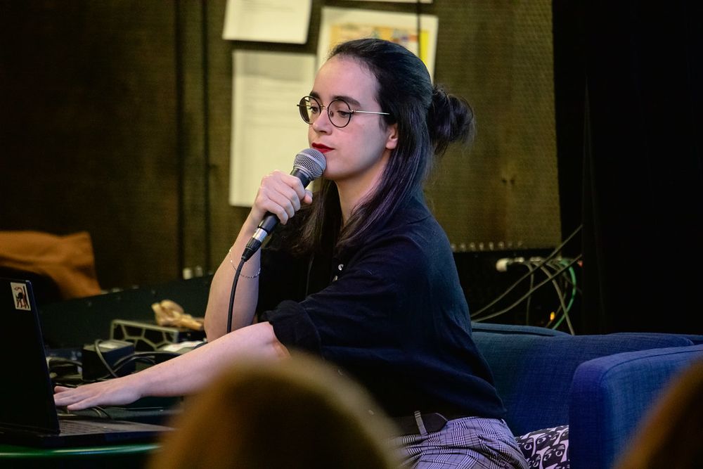 Credits : Alexandre Nachbauer
A pic of me sitting (black hair, round glasses, red lipstick, dressed in blakc and grey)  in an armchair holding a microphone looking down at my computer, ready to skip a slide